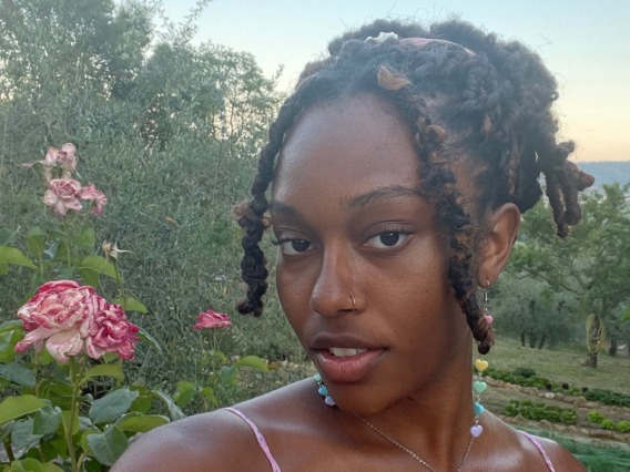 The front desk assistant poses in front of the camera at an angle with a green landscape behind her. Her hair is up with two pieces of hair out in the front, her hair is up, and a pink tank top. 