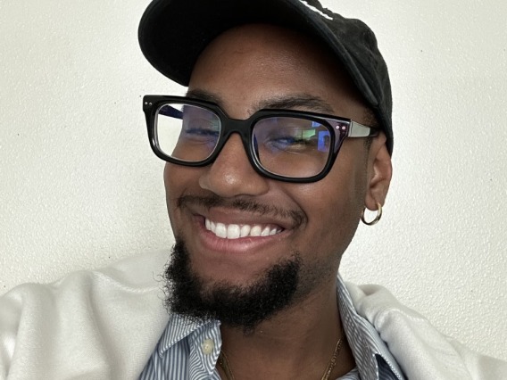 student worker smiling in front of a white wall wearing a black hat, black glasses, blue button up and jacket draped over the shoulders
